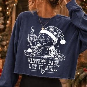 May include: A navy blue long-sleeve cropped sweatshirt featuring a white graphic of a melting snowman wearing a Santa hat and holding a steaming mug. The text reads, "WINTER'S PACE, LET IT MELT." with a small heart icon below the lettering.
