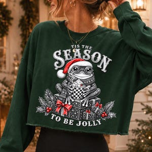 Puede incluir: Una sudadera corta de color verde oscuro con el gr&aacute;fico de una rana con gorro de Pap&aacute; Noel y bufanda, sosteniendo una taza de cacao. El texto 'TIS THE SEASON TO BE JOLLY' rodea a la rana sentada sobre cajas de regalo envueltas.