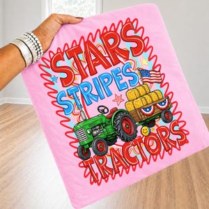 Pode incluir: Cobertor rosa com um trator verde, fardos de feno e uma bandeira americana. O cobertor tem as palavras "Stars Stripes Tractors" em vermelho e azul. O cobertor &eacute; segurado por uma pessoa.