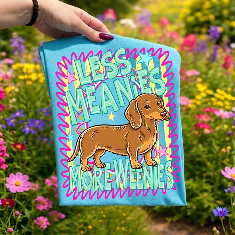 May include: A light blue t-shirt featuring a brown dachshund illustration. The shirt has the text "LESS MEANIES MORE WEENIES" in a playful font with pink and yellow accents. The shirt is held up in front of a colorful flower garden.