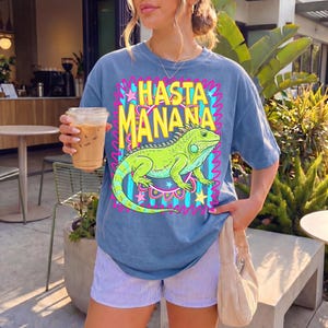 K&ouml;nnte beinhalten: Blaues T-Shirt mit dem gelben Schriftzug "Hasta Ma&ntilde;ana" und einer gr&uuml;nen Leguan-Grafik. Das Design ist mit rosa, blauen und gelben Akzenten umrahmt. Die Person tr&auml;gt blau-wei&szlig; gestreifte Shorts und h&auml;lt ein Kaffeegetr&auml;nk.