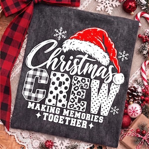 May include: A dark gray t-shirt featuring a white graphic with the text "Christmas CREW" and "MAKING MEMORIES TOGETHER". The design includes a red Santa hat, white snowflakes, and letters filled with buffalo plaid, polka dot, and leopard patterns.