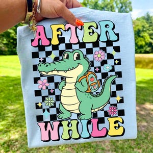 K&ouml;nnte beinhalten: Hellblaues T-Shirt mit einem Cartoon-Alligator mit Rucksack, vor einem schwarz-wei&szlig; karierten Hintergrund. Das Shirt zeigt die Worte "AFTER" und "WHILE" in bunten Buchstaben sowie Blumen- und Sterngrafiken.