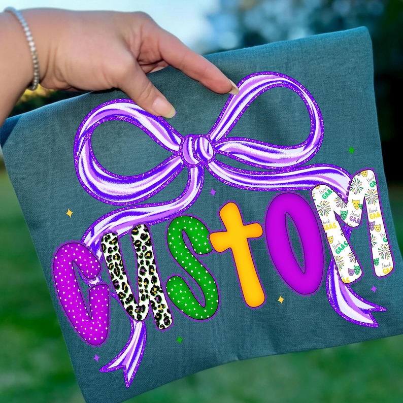 May include: A teal-colored t-shirt featuring the word "CUSTOM" in a playful, colorful design. The letters are in various patterns and colors, including purple polka dots, leopard print, green polka dots, and yellow. A purple bow and ribbons surround the word.