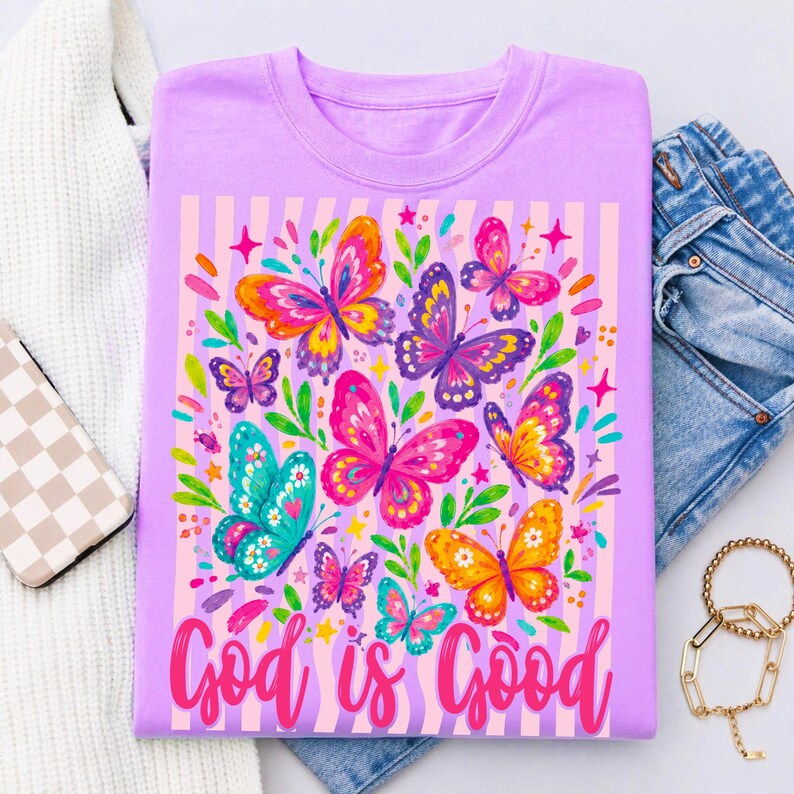 May include: A light purple crew neck t-shirt featuring a colorful graphic of various butterflies in shades of pink, orange, yellow, and teal, set against a background of vertical stripes with the text God is Good in a pink cursive script.