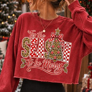 Op de afbeelding: Een rood, langmouwig, kort sweatshirt met een grafische print met de tekst Tis the SEASON to be Merry. Het ontwerp bevat een kerstboom met luipaardprint, een rood-witte strik met stippen, hulstbladeren en rode bessen.