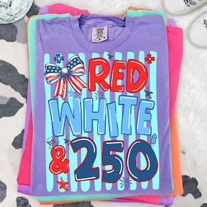 Puede incluir: Una pila de camisetas coloridas. La camiseta superior es morada con las palabras "RED WHITE & 250" en rojo y azul, junto con un lazo y estrellas. Otras camisetas son rosas, naranjas y verdes.