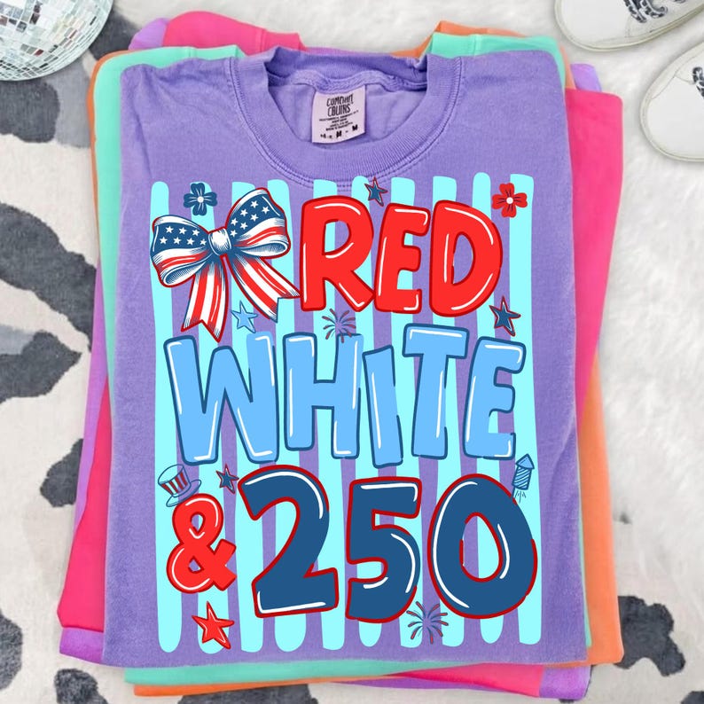 Puede incluir: Una pila de camisetas coloridas. La camiseta superior es morada con las palabras "RED WHITE & 250" en rojo y azul, junto con un lazo y estrellas. Otras camisetas son rosas, naranjas y verdes.