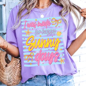 Pu&ograve; includere: T-shirt lilla con la scritta "I was made for happy sunny days" in rosa e giallo. Il design include stelle marine, fiocchi e una conchiglia. La maglietta ha una vestibilit&agrave; ampia.