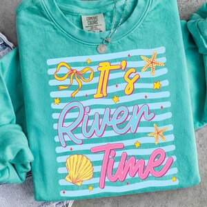 May include: Teal Comfort Colors sweatshirt with the phrase "It's River Time" in yellow, pink, and blue. The design includes a bow, starfish, shell, and stars. A silver necklace is visible.