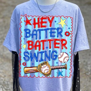 K&ouml;nnte beinhalten: Hellgraues T-Shirt mit Baseball-Motiv. Das Design zeigt die Worte "HEY BATTER BATTER SWING" in Rot und Blau, zusammen mit einem Baseball, Schl&auml;ger und Handschuh. Das T-Shirt hat kurze &Auml;rmel.