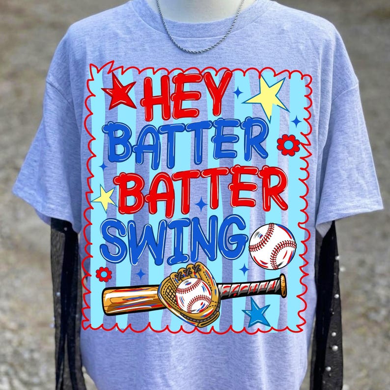 K&ouml;nnte beinhalten: Hellgraues T-Shirt mit Baseball-Motiv. Das Design zeigt die Worte "HEY BATTER BATTER SWING" in Rot und Blau, zusammen mit einem Baseball, Schl&auml;ger und Handschuh. Das T-Shirt hat kurze &Auml;rmel.