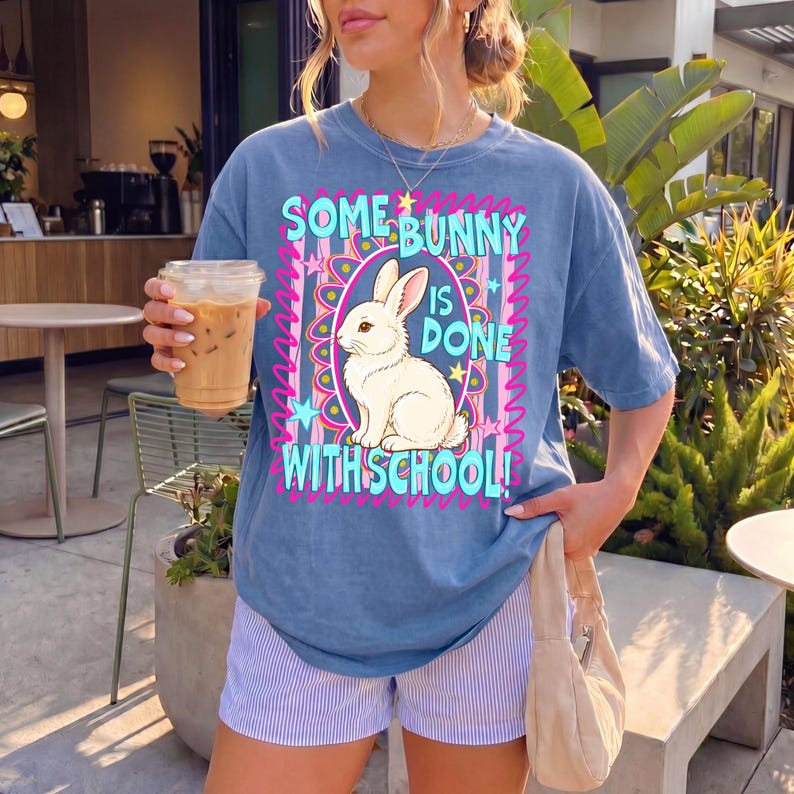 Może przedstawiać: Niebieska koszulka z napisem "SOME BUNNY IS DONE WITH SCHOOL!" i grafiką białego kr&oacute;lika. Koszulka ma kolorowy design z r&oacute;żowymi, niebieskimi i ż&oacute;łtymi akcentami. Osoba ma na sobie niebiesko-białe szorty w paski.