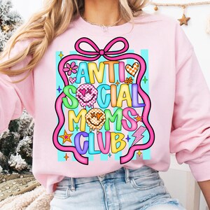 K&ouml;nnte beinhalten: Hellrosa Sweatshirt mit dem Schriftzug "ANTI SOCIAL MOMS CLUB" in einem farbenfrohen, cartoonartigen Design. Das Design umfasst Blumen, Sterne und Blitze. Das Sweatshirt hat lange &Auml;rmel und einen Rundhalsausschnitt.