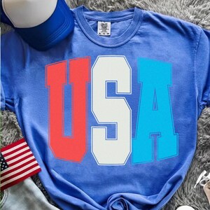 May include: A blue t-shirt with the word "USA" in red, white, and blue block letters. The shirt is a crew neck style. A blue and white baseball cap and an American flag are also visible.