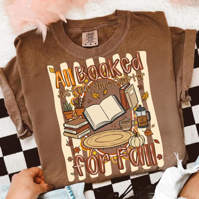K&ouml;nnte beinhalten: Ein braunes Kurzarm-T-Shirt mit einem Grafikdruck und dem Text All Booked Just One More Chapter for Fall. Das Design zeigt ein aufgeschlagenes Buch, einen B&uuml;cherstapel, eine Kaffeetasse, eine French Press, einen K&uuml;rbis und Topfpflanzen.