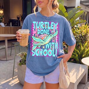 以下が含まれることがあります： 「TURTLEY DONE WITH SCHOOL！」の文字とウミガメのグラフィックがプリントされた青いTシャツ。ストライプのショートパンツとベージュのハンドバッグを合わせています。透明なカップに入ったアイスコーヒーを持っています。