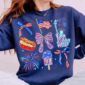 K&ouml;nnte beinhalten: Marineblaues Sweatshirt mit patriotischen Grafiken, darunter die Freiheitsstatue, eine amerikanische Flagge, Schleifen, Feuerwerk, ein Hotdog und ein Eis am Stiel. Das Design verwendet Rot-, Wei&szlig;- und Blaut&ouml;ne.