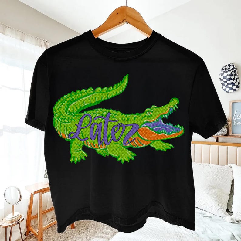 Pu&ograve; includere: T-shirt nera con un disegno di alligatore verde e la scritta "Lātez" in viola. L'alligatore ha dettagli arancioni e viola. La maglietta &egrave; appesa a un appendiabiti di legno.