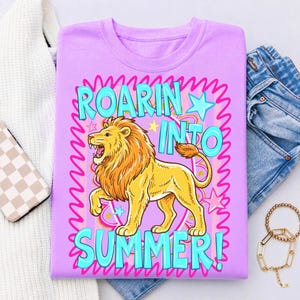 May include: A lilac-colored t-shirt with a cartoon lion graphic. The lion is yellow with an orange mane and is roaring. The text "ROARIN' INTO SUMMER!" is in turquoise and pink. The shirt is folded with jeans and a sweater.