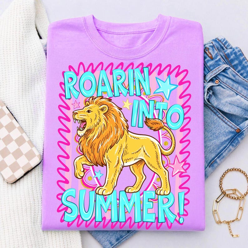 May include: A lilac-colored t-shirt with a cartoon lion graphic. The lion is yellow with an orange mane and is roaring. The text "ROARIN' INTO SUMMER!" is in turquoise and pink. The shirt is folded with jeans and a sweater.