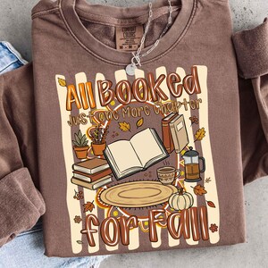 K&ouml;nnte beinhalten: Ein braunes Langarm-Sweatshirt mit einem Grafikdruck und dem Text "All Booked Just one More Chapter for Fall". Das Design zeigt Illustrationen eines offenen Buches, eines B&uuml;cherstapels, einer Kaffeetasse, einer Kaffeepresse und eines K&uuml;rbisses.