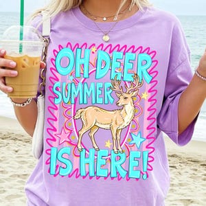 K&ouml;nnte beinhalten: Ein lila T-Shirt mit dem Text "OH DEER SUMMER IS HERE!" in T&uuml;rkis, Pink und Gelb. Eine hellbraune Hirschgrafik befindet sich in der Mitte des Textes. Das T-Shirt hat kurze &Auml;rmel.