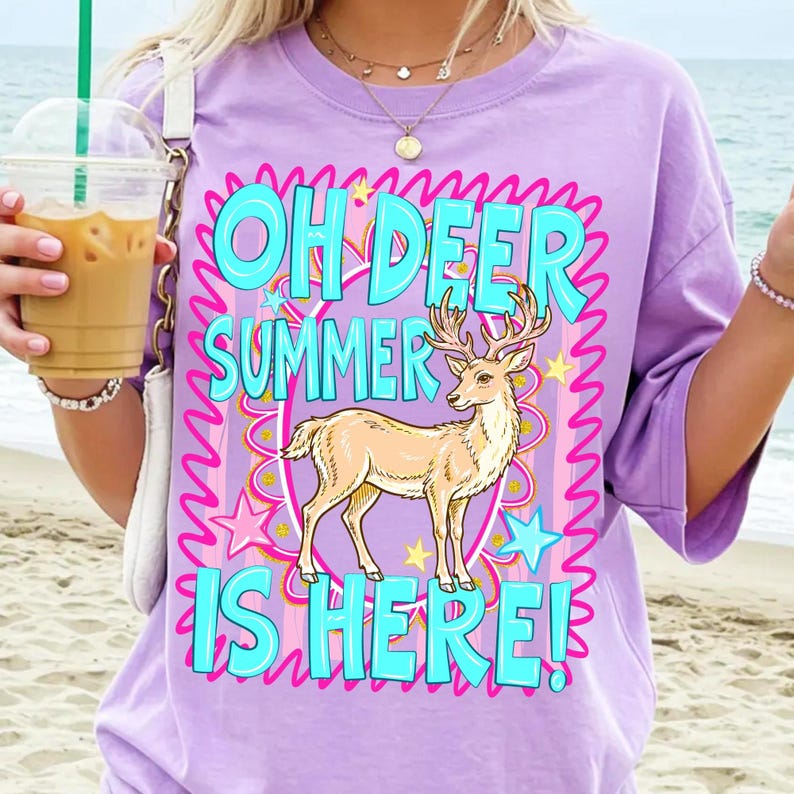 K&ouml;nnte beinhalten: Ein lila T-Shirt mit dem Text "OH DEER SUMMER IS HERE!" in T&uuml;rkis, Pink und Gelb. Eine hellbraune Hirschgrafik befindet sich in der Mitte des Textes. Das T-Shirt hat kurze &Auml;rmel.