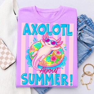 Peut inclure: T-shirt lilas avec un dessin d'axolotl portant des lunettes de soleil et flottant sur une bou&eacute;e fleurie. Le texte "AXOLOTL ABOUT SUMMER!" est en bleu. Le t-shirt est pli&eacute; sur une surface blanche.