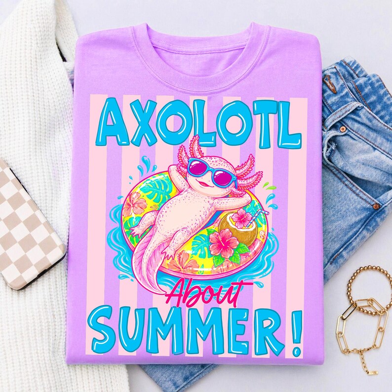 Peut inclure: T-shirt lilas avec un dessin d'axolotl portant des lunettes de soleil et flottant sur une bou&eacute;e fleurie. Le texte "AXOLOTL ABOUT SUMMER!" est en bleu. Le t-shirt est pli&eacute; sur une surface blanche.