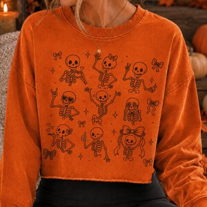 Op de afbeelding: Een oranje sweater met lange mouwen en ronde hals, voorzien van een zwarte grafische print van meerdere skeletten in diverse houdingen, zoals zwaaiend, hartjes vormend en met strikjes, afgewisseld met kleine ster- en striksymbolen.