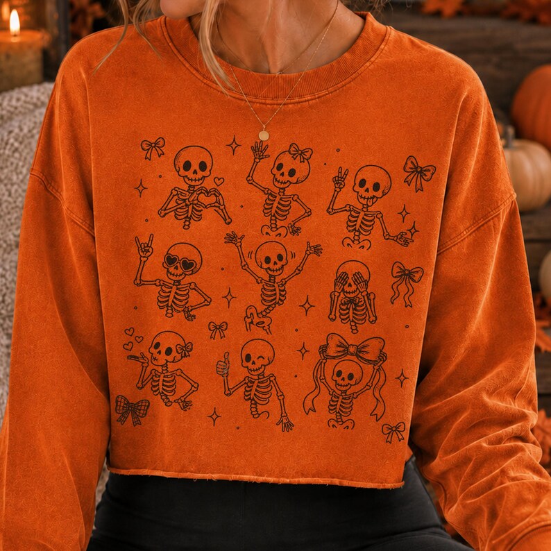 Op de afbeelding: Een oranje sweater met lange mouwen en ronde hals, voorzien van een zwarte grafische print van meerdere skeletten in diverse houdingen, zoals zwaaiend, hartjes vormend en met strikjes, afgewisseld met kleine ster- en striksymbolen.
