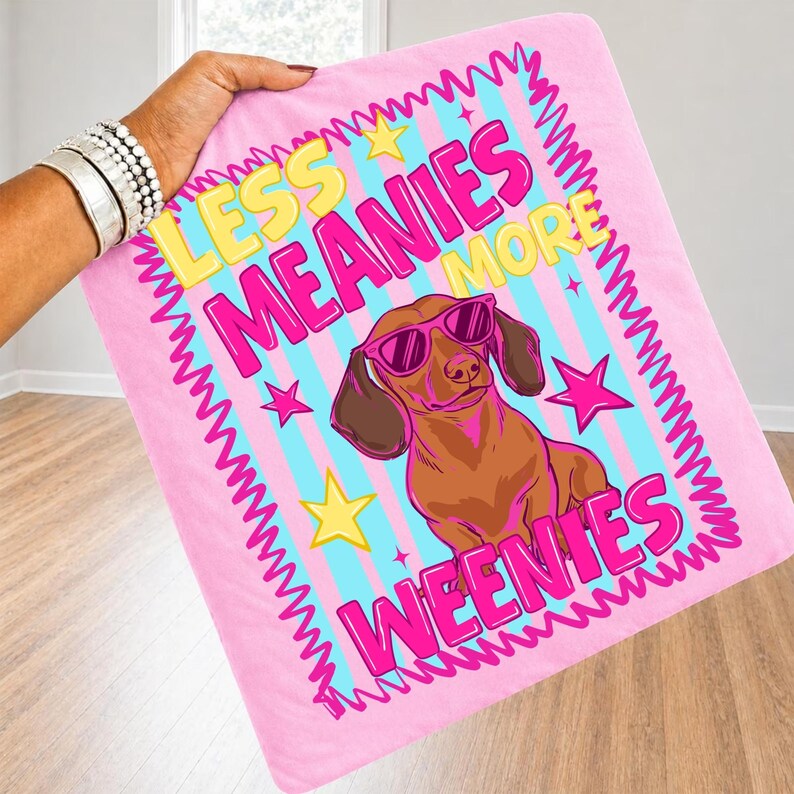 May include: Pink blanket featuring the text "LESS MEANIES MORE WEENIES" and a cartoon dachshund wearing sunglasses. The blanket has a pink zig-zag border, yellow stars, and blue and pink stripes.
