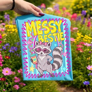 以下が含まれることがあります： ハート型のサングラスとピンクのリボンをつけた漫画のキャラクターのアライグマが描かれた、水色のTシャツ。「Messy Bestie」の文字はピンクの縁取りのある黄色です。アライグマはハートのマークのついた飲み物を持っています。シャツにはピンクの波型のボーダーがあります。