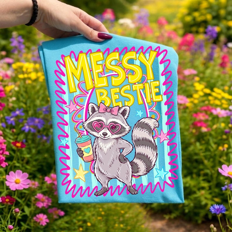 以下が含まれることがあります： ハート型のサングラスとピンクのリボンをつけた漫画のキャラクターのアライグマが描かれた、水色のTシャツ。「Messy Bestie」の文字はピンクの縁取りのある黄色です。アライグマはハートのマークのついた飲み物を持っています。シャツにはピンクの波型のボーダーがあります。
