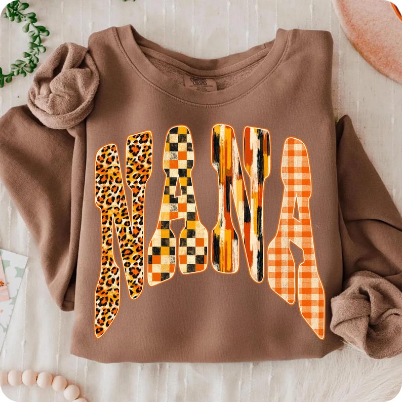 May include: A brown long-sleeve sweatshirt featuring the word NANA printed in large, stylized letters. Each letter has a unique pattern: the first N is leopard print, the A is a black and orange checkerboard, the second N is orange brushstrokes, and the final A is an orange gingham plaid.