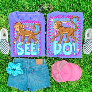 May include: A flat lay of a casual outfit on green grass. The outfit includes two purple t-shirts with a monkey graphic and the words "SEE DO!", denim shorts, a pink trucker hat, a blue scrunchie, and checkered slip-on sneakers.
