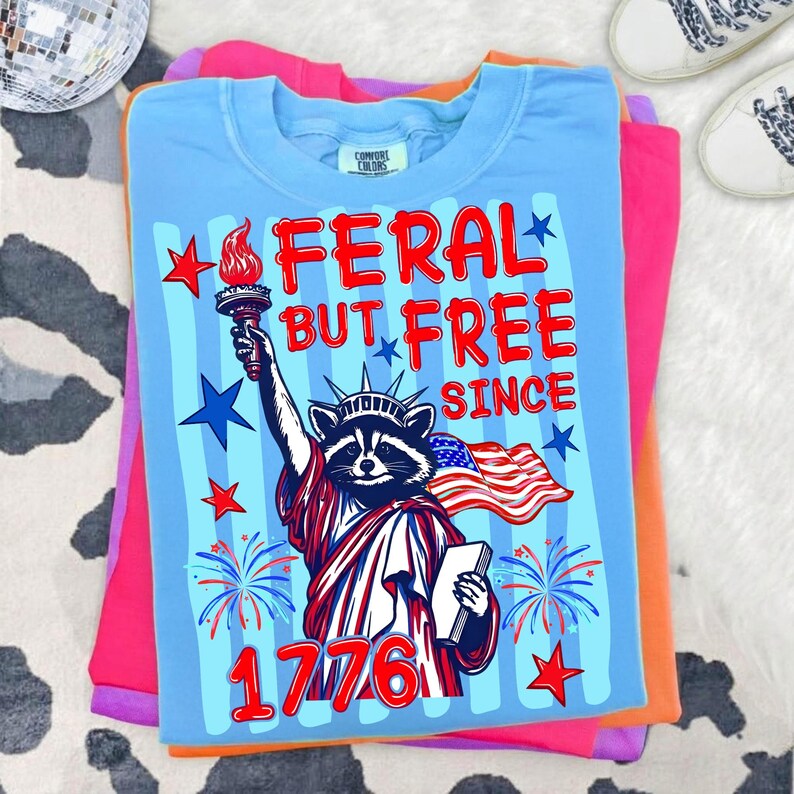 May include: A light blue t-shirt with the text "FERAL BUT FREE SINCE 1776" and a graphic of a raccoon as the Statue of Liberty holding a torch and the American flag. The shirt is folded on top of other colorful shirts.