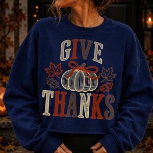 Puede incluir: Una sudadera azul marino de manga larga con un estampado gr&aacute;fico que incluye el texto GIVE THANKS en una fuente de l&iacute;neas m&uacute;ltiples. El dise&ntilde;o presenta una calabaza central de l&iacute;neas blancas con un lazo rojo y hojas de arce y roble.
