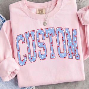 Op de afbeelding: Een lichtroze sweatshirt met het woord "CUSTOM" in grote, blauwe letters gevuld met witte en rode sterren. De sweatshirt heeft een ronde hals en lange mouwen. Een zilveren ketting met een ronde hanger is zichtbaar.