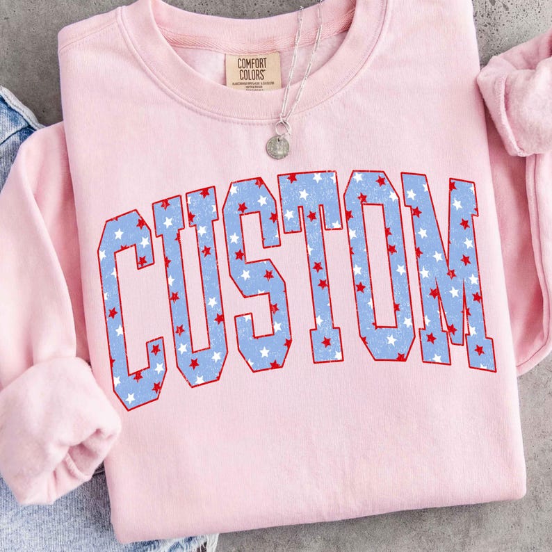 Op de afbeelding: Een lichtroze sweatshirt met het woord "CUSTOM" in grote, blauwe letters gevuld met witte en rode sterren. De sweatshirt heeft een ronde hals en lange mouwen. Een zilveren ketting met een ronde hanger is zichtbaar.