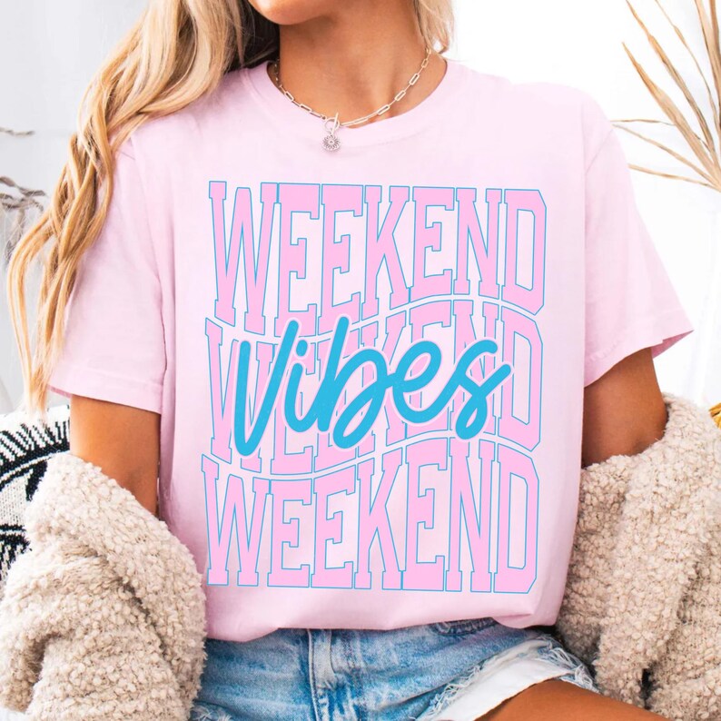 K&ouml;nnte beinhalten: Hellrosa T-Shirt mit dem Schriftzug "WEEKEND VIBES" in einem gestapelten Design. Der Text ist in hellblauer Schrift gehalten und mit dunklerem Rosa umrandet. Das T-Shirt hat kurze &Auml;rmel und einen Rundhalsausschnitt.