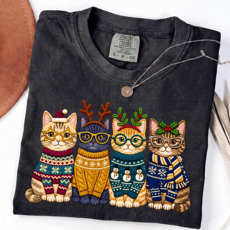 May include: A charcoal gray t-shirt featuring a graphic of four cats wearing holiday sweaters, glasses, and festive accessories like reindeer antlers, a Santa hat, and holly. The shirt has a Comfort Colors brand label at the inner neckline.