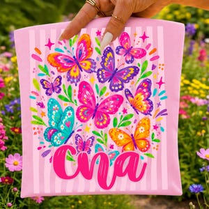 May include: A light pink fabric pouch featuring a vertical striped pattern and a colorful graphic print of various butterflies in shades of pink, purple, orange, and teal, with the name "Cna" written in a bold, cursive, bright pink script font.