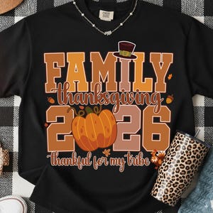 Pode incluir: Uma t-shirt preta de manga curta com uma estampa gr&aacute;fica contendo o texto FAMILY THANKSGIVING 2026 e thankful for my tribe. O design inclui uma ab&oacute;bora laranja grande, um chap&eacute;u de peregrino castanho, bolotas e um peru em tons de outono.