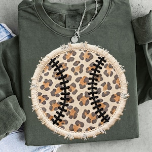 Puede incluir: Sudadera verde oliva con diseño de béisbol. El béisbol tiene un estampado de leopardo y costuras negras. La sudadera tiene mangas largas y cuello redondo. Se ve un collar plateado.