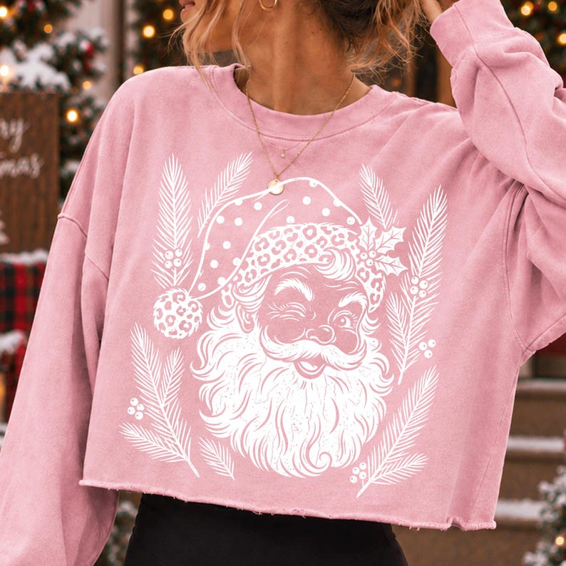 Puede incluir: Una sudadera corta de color rosa claro con un estampado gr&aacute;fico blanco de un Santa Claus gui&ntilde;ando un ojo, que lleva un gorro con estampado de leopardo. El dise&ntilde;o est&aacute; rodeado por ilustraciones blancas de ramas de pino y bayas de acebo.
