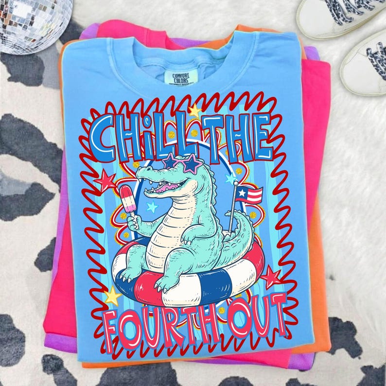 Puede incluir: Camiseta azul claro con un dise&ntilde;o de un caim&aacute;n con gafas de sol flotando en una boya roja, blanca y azul. El texto "CHILL THE FOURTH OUT" est&aacute; en la camiseta. La camiseta est&aacute; doblada sobre otras camisetas coloridas.