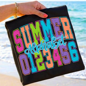 May include: Black t-shirt with the words "SUMMER Alphabet 0123456" in bright, colorful lettering. The letters are outlined in a contrasting color, with a beach and ocean background.