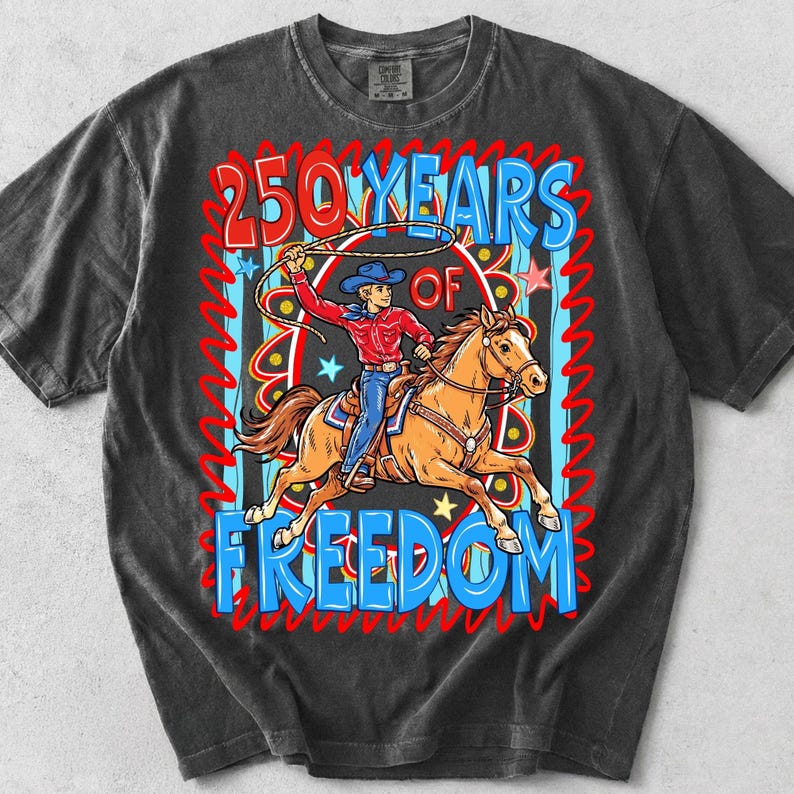 Puede incluir: Camiseta gris oscuro con un gr&aacute;fico colorido de un vaquero a caballo, lazando. El texto dice "250 YEARS OF FREEDOM" en rojo y azul. El vaquero lleva una camisa roja, vaqueros azules y un sombrero azul.